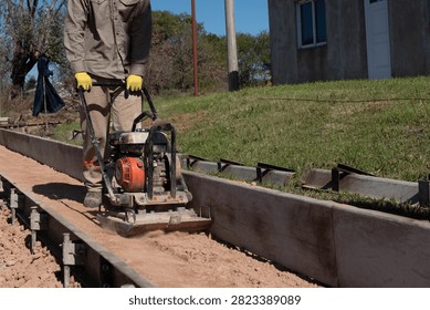 Worker compacting soil subbase with plate compactor between metal formwork for curb construction