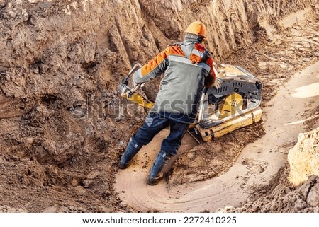 A worker compacts soil or sand with a vibrating plate in a trench at a construction site close-up. Vibratory soil compaction for laying underground utilities