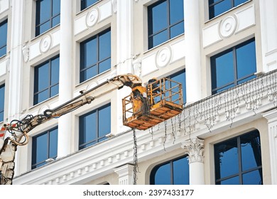 Worker in cradle decorating building facade, install Christmas lights. Electrician on aerial platform installing Christmas lights ornaments. Man hanging festive garlands on building