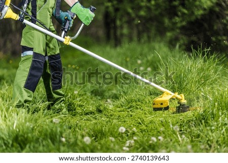 Worker cuts grass in a field using a string trimmer on a sunny day.