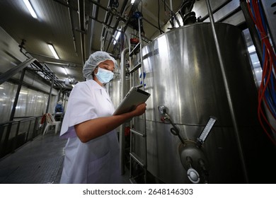 A worker in a dairy processing plant checks machinery and records data on a tablet while wearing protective gear. This routine activity ensures compliance with safety standards and efficiency.