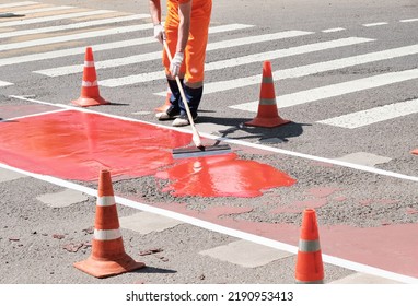 Worker Draws Line At Crosswalk. Traffic Cones With Orange And White Stripes In Background, Standing On Asphalt During Road Construction Work.