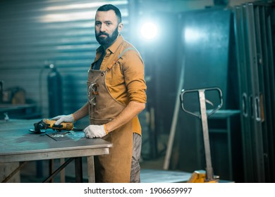 Worker dressed in apron doing some metalwork, grinding steel product at the factory. Handsome artisan at the workshop of metal products. High quality photo