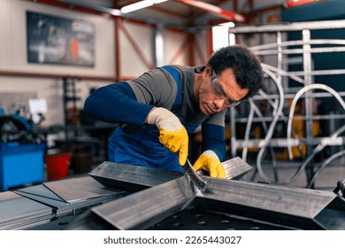 A worker dressed in overalls and protective gloves, wearing safety glasses, fixes a metal object on the workbench very carefully