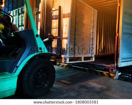 Worker driving forklift loading shipment carton boxes goods on wooden pallet at loading dock from container truck to warehouse cargo storage in freight logistics, transportation industrial, delivery