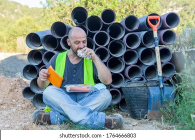 Worker Eating A Sandwich In Reflective Vest Sitting In The Shade At Lunch Time Leaning On A Stack Of Construction Drain Pipes. Work Lifestyle.