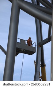 Worker in elevated work platform using exothermic cutting torch to dismantle large metal structure, wearing protective safety gear including hard hat and protective suit.