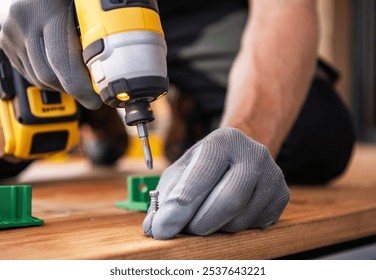 A worker is focused on using a cordless power drill to securely fasten screws into a wooden surface while wearing gloves.