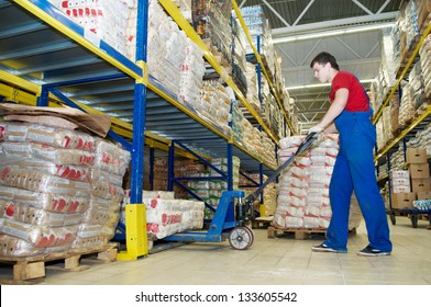 Worker With Fork Pallet Truck Stacker In Warehouse Loading Group Of Food Packages