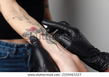 Worker in gloves applying cream on woman's arm with tattoo against light background, closeup