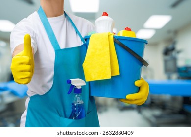 A worker in gloves displays a thumbs up while holding a cleaning bucket filled with supplies. The clean environment of a medical facility enhances hygiene and safety standards.