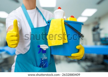 A worker in gloves displays a thumbs up while holding a cleaning bucket filled with supplies. The clean environment of a medical facility enhances hygiene and safety standards.