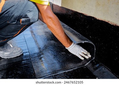 Worker in gloves prepare for melting, manually place and unroll rolled up, sheet of waterproofing installation over concrete. Residential building under construction placing water insulation