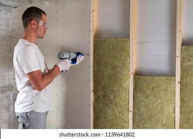 Worker in goggles with screwdriver working on insulation. Drywall on wall beams, insulating rock wool staff in wooden frame. Comfortable warm home, economy, construction and renovation concept.