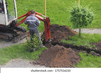 A worker in gray overalls stands next to a working compact rubber tracked excavator, which carefully digs a ditch in the Park with a bucket between bushes and paving stones on summer day, landscaping