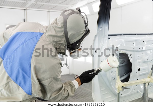 a worker\
in a gray protective suit are painting car door parts  spray.  hard\
work. mask and protective\
shield.