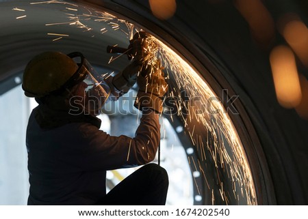 The worker is grinding steel material and weld metal with hand grinding machine from inside of large pipe. Grinding is an abrasive machining process that uses a grinding wheel as the cutting tool.