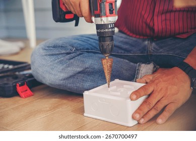 Worker hand using an electric drill with step drill bit to make a hole in a white plastic box, concept of DIY, construction, repair, and electrical installation.