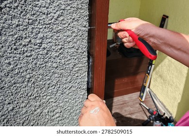 ​Close-up of worker hands using a manual screwdriver to fix a wooden frame to a house wall during renovation works, with a spirit level tool in the background.