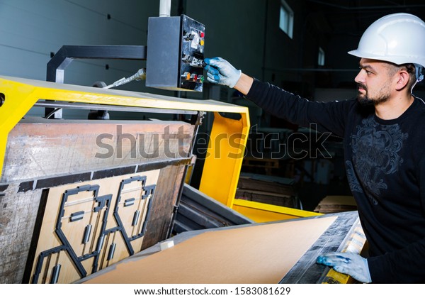 Worker in a hard hat\
turning on a punching machine. Cardboard boxes factory. Paper die\
cutting machine