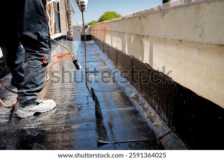 Worker is heating, melting bitumen waterproofing installation over concrete using propane blowtorch, spreading with working tool. Residential building under construction placing water insulation