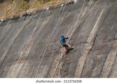 Worker At Heights. Worker With No Safety Helmet Is Working And Fixing Landslide Safety Net 