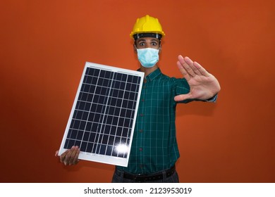 Worker With Helmet And Pandemic Mask Holding A Solar Photovoltaic Panel Isolated. Solar Worker Isolated In Orange Background.
