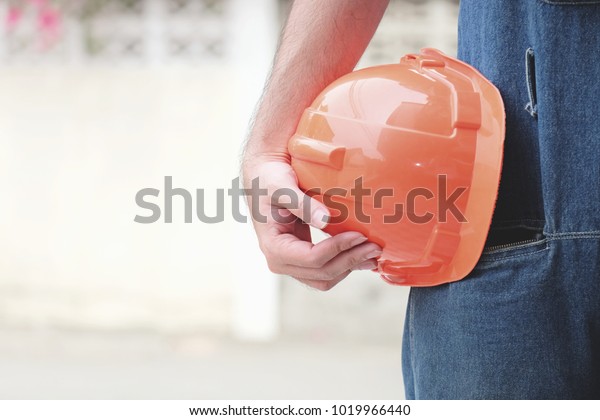 Worker holding with\
orange plastic helmet in hand, Mechanic in jeans with helmet in\
hand while ralax time