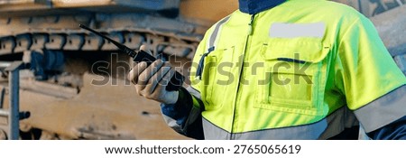 Worker holding radio communicator while supervising heavy machinery.