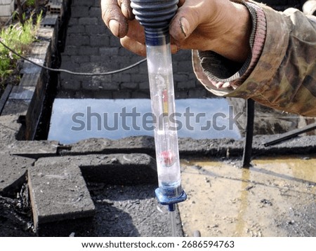 A worker holds a hydrometer or densimeter to test the specific gravity of a liquid, with a small pit or foundation filled with water in the background.