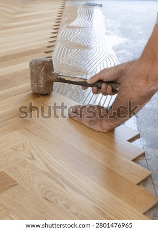 Worker installing herringbone parquet flooring with rubber mallet during hardwood floor installation in apartment renovation. Carpenter laying wooden parquet planks over adhesive
