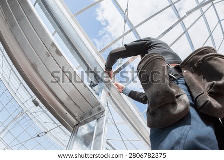 Worker installing a metal frame on a glass greenhouse structure, low angle view
