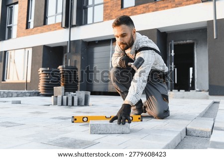 Worker installing paving bricks on an outdoor patio using a spirit level and rubber hammer at a modern building