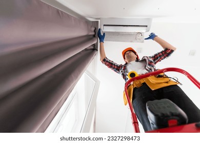 Worker installing or repairing air conditioner. Young man in uniform standing on ladder with toolbox and installing new air conditioner on wall. AC installation, maintenance and repair service concept - Powered by Shutterstock