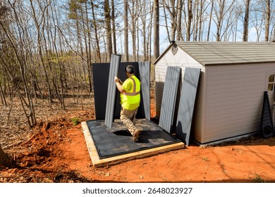 Worker installs large panels while standing on wooden platform during assembling storage shed surrounded by trees.