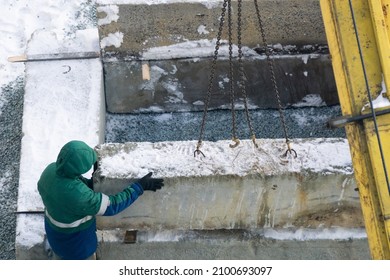 A Worker Installs A Snow-covered Foundation Block Using A Manipulator Crane. Migrant Labor In Winter Conditions, Construction Work.