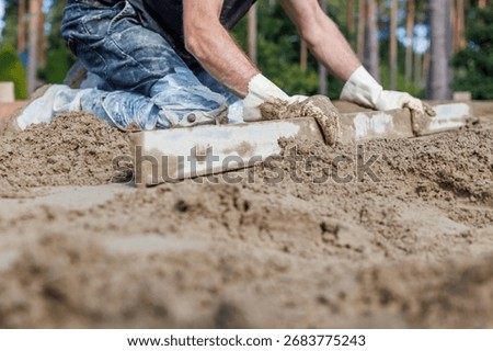 A worker kneels while leveling sand with a straightedge tool, focusing on precision. Tall trees in the background suggest an outdoor environment.