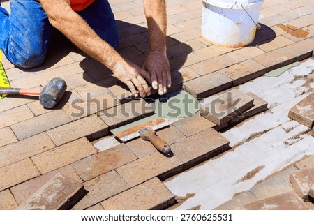 Worker is laying bricks on ground in patio area while using tools for installation.