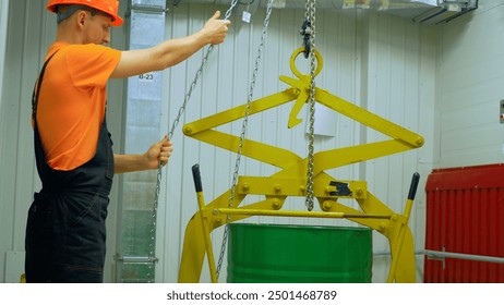 Worker lifts chemical waste barrel with chain hoist in industrial plant. Emphasizing safety protocols, protective gear. Chemical waste handling requires precision, caution. Worker ensures secure. - Powered by Shutterstock