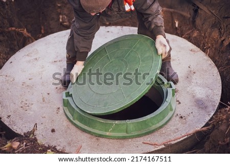 A worker lifts the manhole cover of a sewer well. Construction and maintenance of septic tanks in the village.