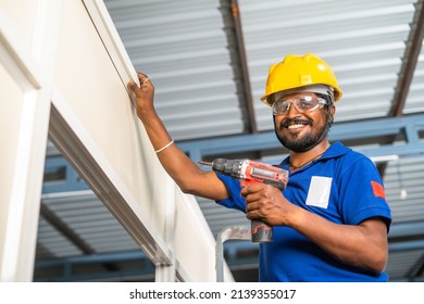 Worker Looking Camera While Fixing Screw Using Drilling Machine Or Gun On Aluminium Partition Frame At Construction Site - Concept Of Maintenance Service, Blue Collar Jobs And Safety.