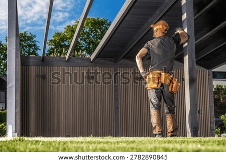 A worker looks at a structure he is building at a construction site. It is day, and the sun shines brightly. The worker wears a cap and has tools in his belt. There are trees in the background.