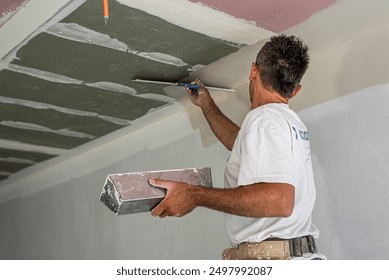 The worker make a plasterboard ceiling. He does taping plaster drywall ceiling joints. He is using special stainless steel taping knife.