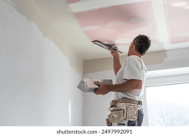 The worker make a plasterboard ceiling. He does taping plaster drywall ceiling joints. He is using special stainless steel taping knife.