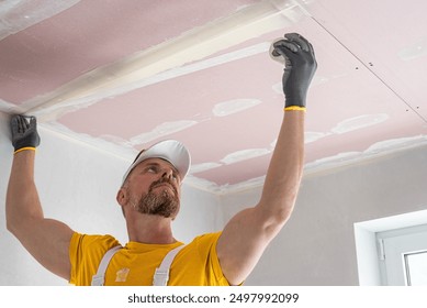 The worker make a plasterboard ceiling. He does taping plaster drywall ceiling joints. He is  putting fiberglass tape into the plaster putty. 