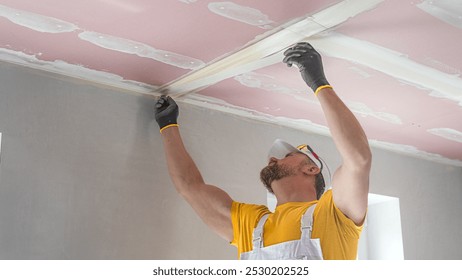 The worker make a plasterboard ceiling. He does taping plaster drywall ceiling joints. He is inserting the glass tape into the putty.