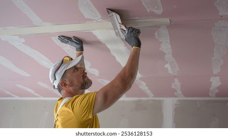 The worker make a plasterboard ceiling. He does taping plaster drywall ceiling joints. He is using special drywall taping knife.