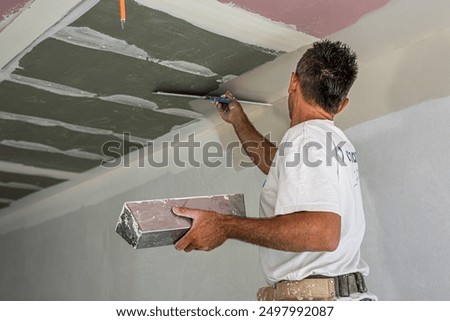 The worker make a plasterboard ceiling. He does taping plaster drywall ceiling joints. He is using special stainless steel taping knife.