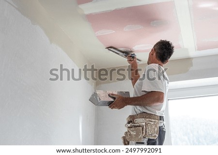 The worker make a plasterboard ceiling. He does taping plaster drywall ceiling joints. He is using special stainless steel taping knife.