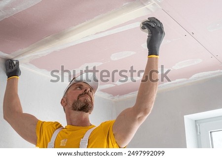 The worker make a plasterboard ceiling. He does taping plaster drywall ceiling joints. He is  putting fiberglass tape into the plaster putty. 